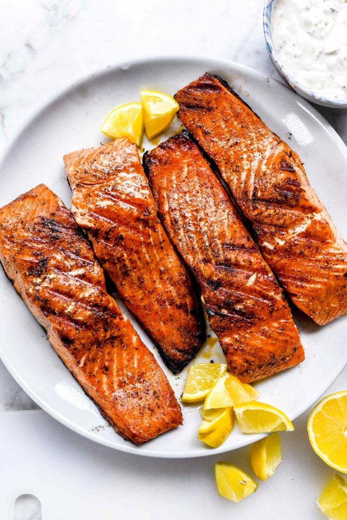 Four grilled salmon fillets on a white plate, with lemon